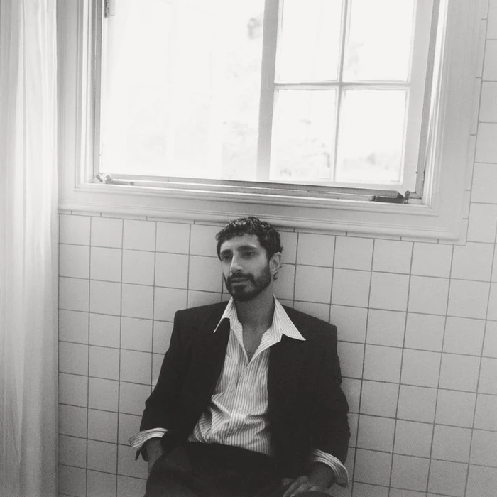 Portrait of Riz Ahmed in a dark suit jacket and striped shirt seated against white tiled wall.