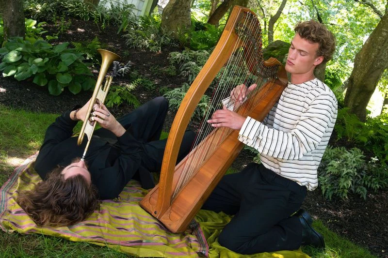 Man playing harp kneeling beside another man lying on a blanket playing trumpet outdoors.