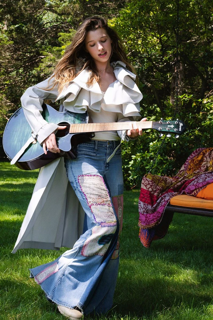 Young woman playing an acoustic guitar outdoors in a garden, wearing patchwork denim jeans and a ruffled white coat.