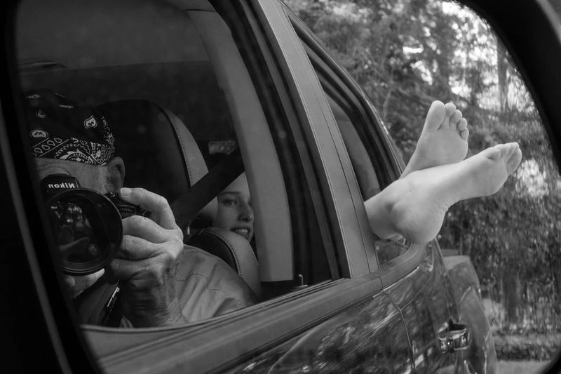 Black and white image of a photographer in a car with bare feet resting out the open window and a passenger visible in the back seat.