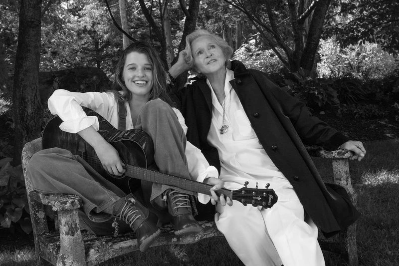 Black and white portrait of a young woman holding a guitar seated next to an older woman on a garden bench outdoors.