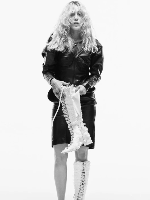 Miley Cyrus holding white lace-up boots wearing a black leather jacket and skirt against a white background