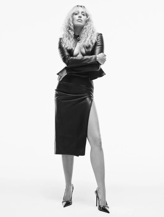 Miley Cyrus wearing a black leather jacket and matching skirt with stilettos against a plain white background