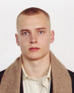 Portrait of a man with short buzzed hair wearing a cream shirt and brown scarf against a plain white background