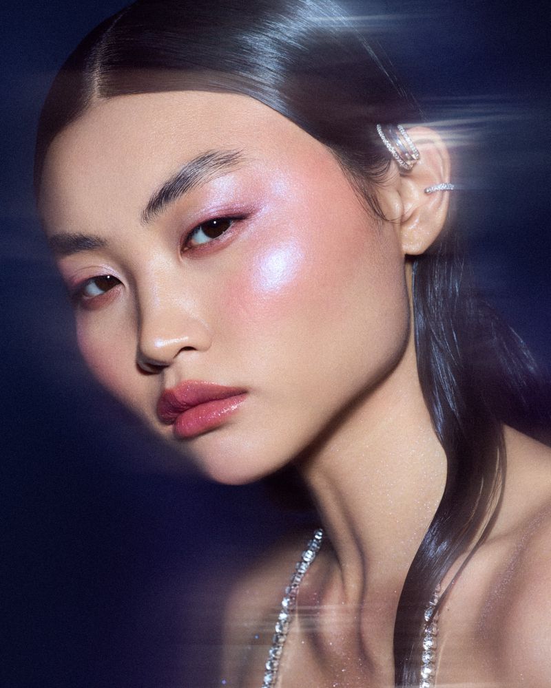 Close-up studio shot of a young Asian woman with pink eye shadow and glossy lips, wearing a sparkly necklace. Dark blue background.