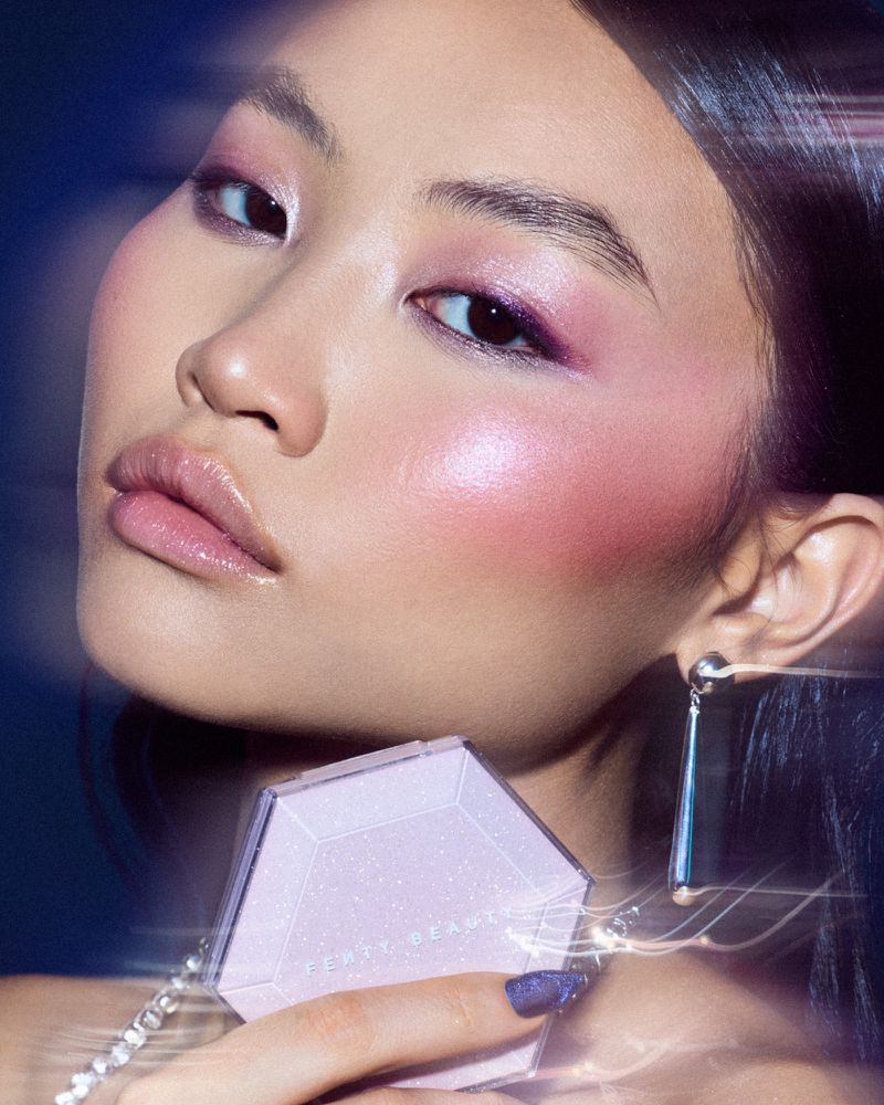 Close-up of a woman with pink eyeshadow holding a light-purple makeup compact with a silver chain and matching nail polish.