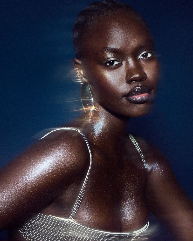 Close-up shot of an African-American woman with dark skin and glossy lips wearing an embellished top with spaghetti straps against a blue background.