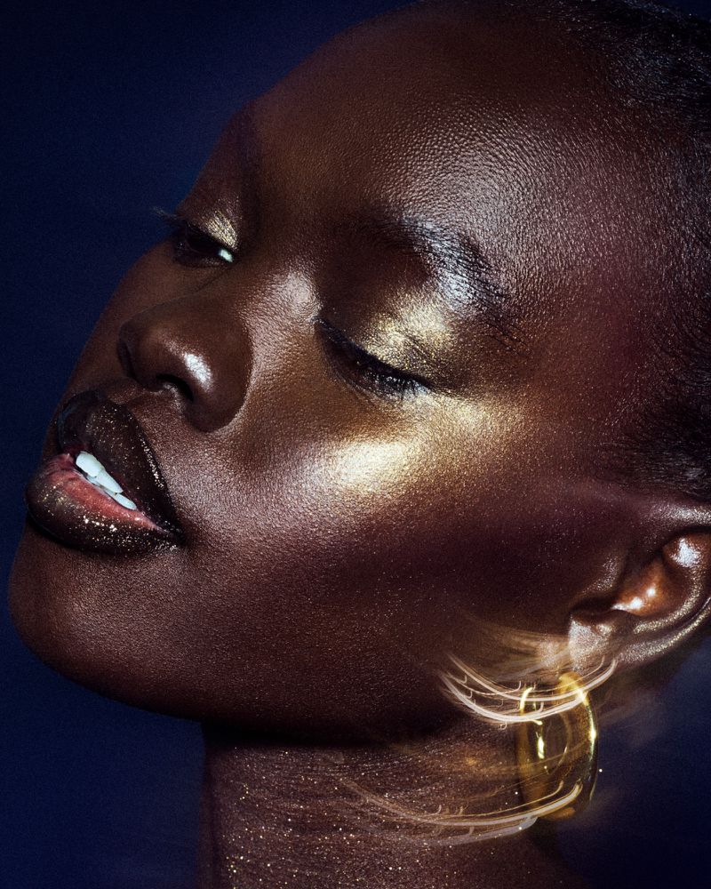 Close-up portrait of a Black woman with closed eyes and glowing skin. Golden jewelry detail accents the look on a dark background.