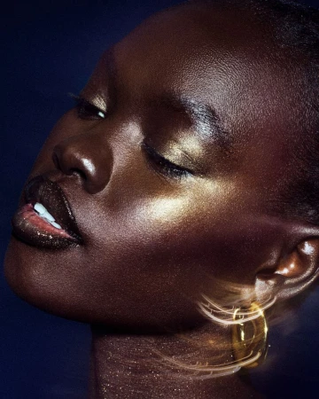 Close-up portrait of a Black woman with closed eyes and glowing skin. Golden jewelry detail accents the look on a dark background.