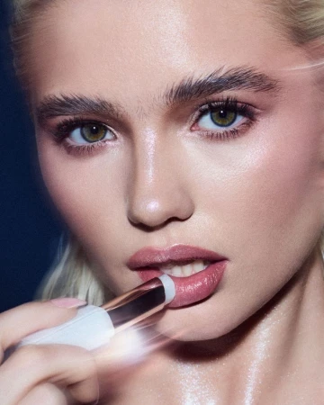 Close-up beauty portrait of a blonde woman applying a lip product from a white and metallic tube against a dark backdrop.