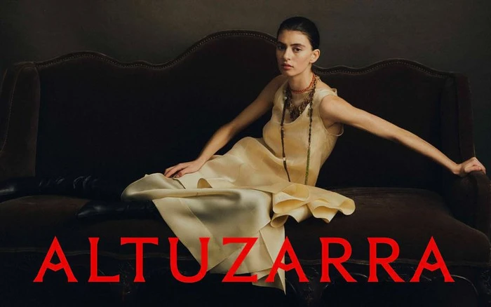 Model wearing pale yellow dress and layered necklaces reclining on a dark velvet sofa