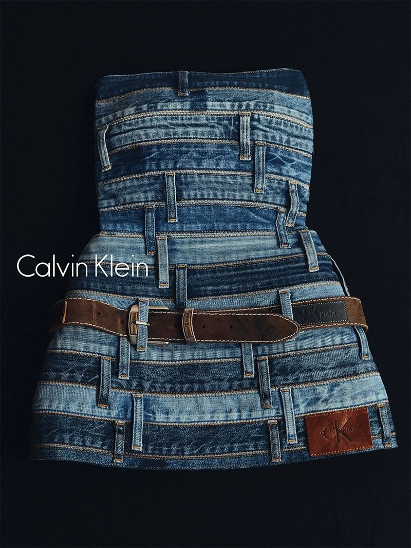 An artistic strapless dress constructed from layered blue denim waistbands and a brown leather belt against a dark background
