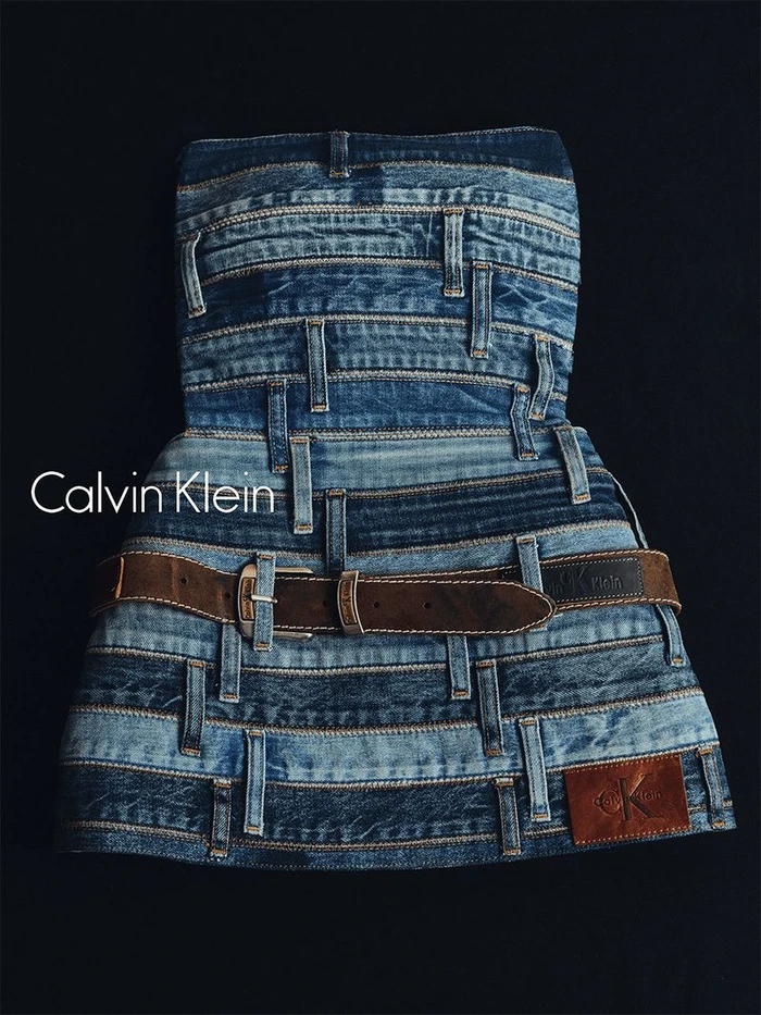 An artistic strapless dress constructed from layered blue denim waistbands and a brown leather belt against a dark background