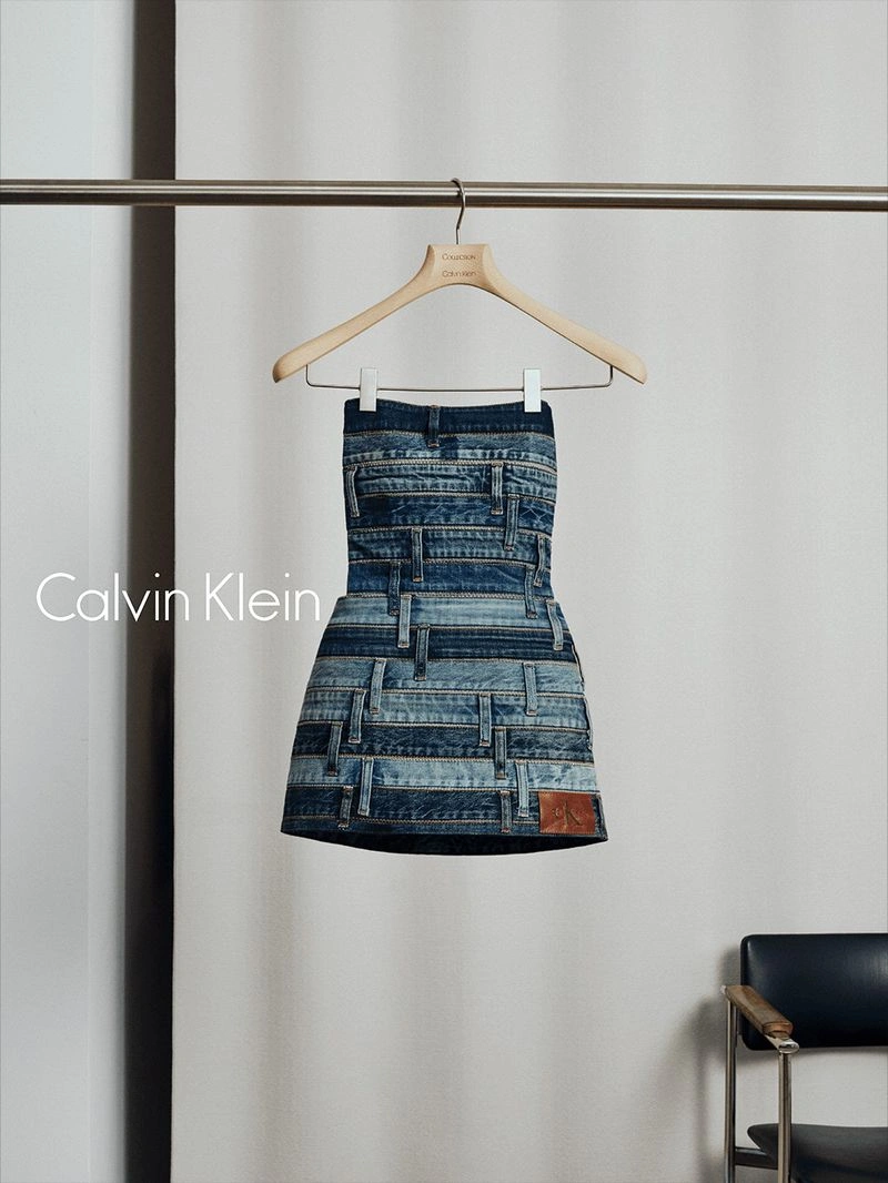 Upcycled denim strapless dress hanging on a wooden hanger against a plain light wall with Calvin Klein text visible.