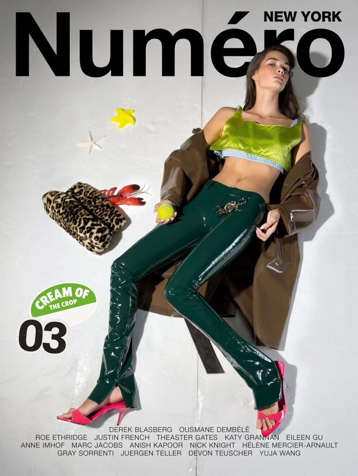 Model lying on a white seamless backdrop wearing green vinyl pants, a shiny green top, and a brown coat, surrounded by props.