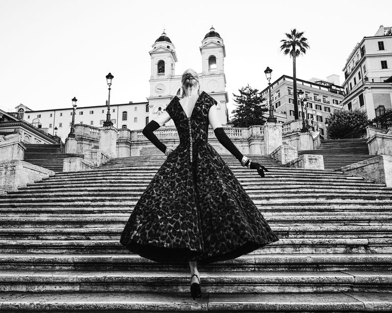 A woman in an elegant dress poses on stone steps in an urban setting, showcasing vintage fashion and architectural surroundings in black and white.