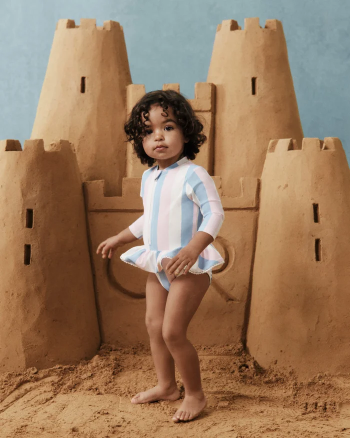 A toddler standing on sand in front of a large sandcastle wearing a pastel striped swimsuit