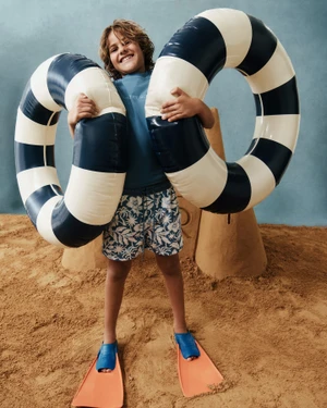 Young boy holding a large black and white striped pool float, wearing blue swim trunks and orange swim fins in the sand.