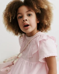 Young child with curly hair wearing a pink dress and carrying a pink bag against a plain background