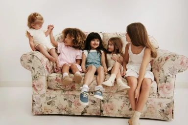 Five young girls seated on a floral patterned sofa against a plain white studio background.