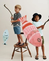 Two young boys holding stick fishing rods with paper cutout fish toys against a plain white studio background.