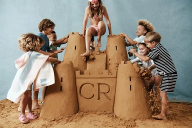 Children playing around a large sandcastle built indoors on a sandy floor with a textured blue backdrop.