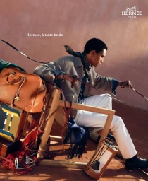 Man seated on wooden chair holding reins outdoors with Hermes travel bag and accessories