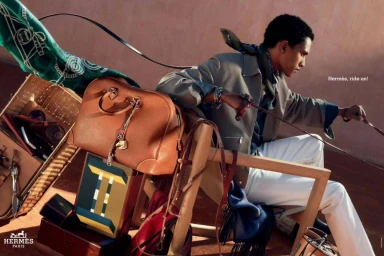 Man wearing tan jacket and white trousers holding leather reins in Hermes Ride On campaign against a warm textured backdrop.