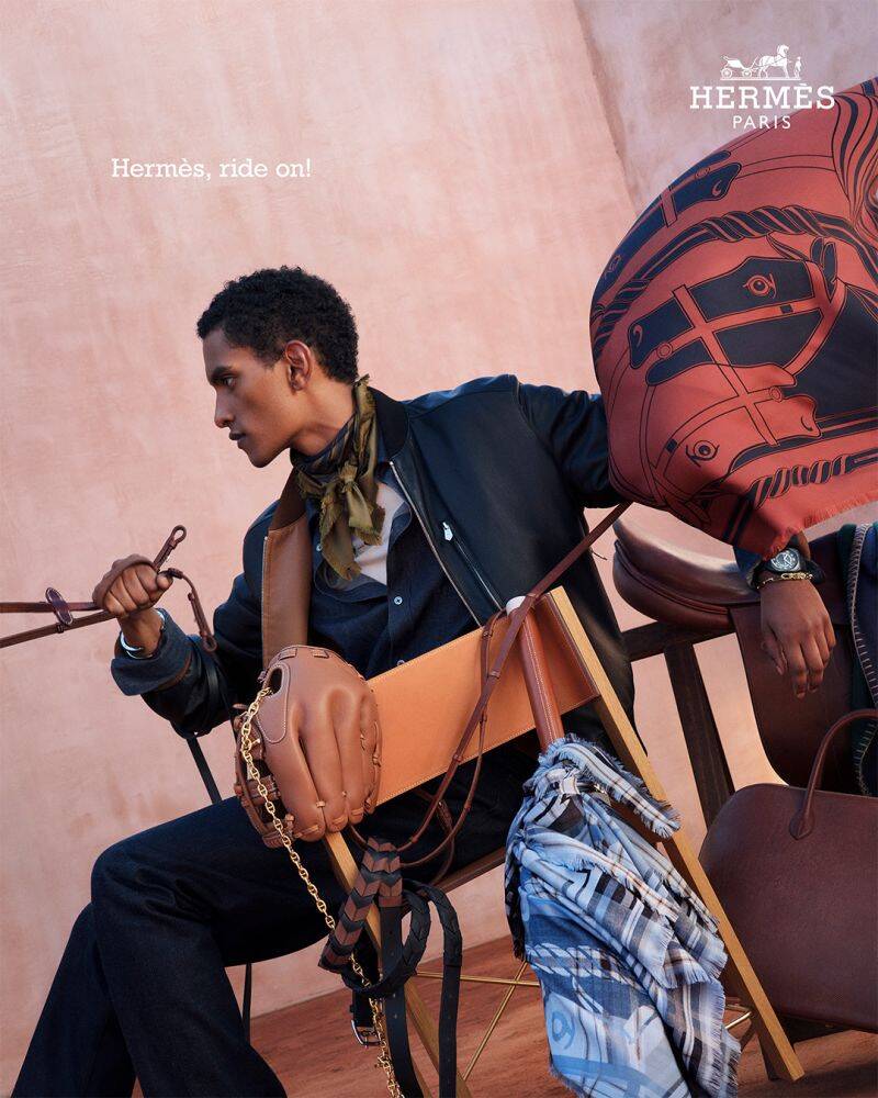 Man seated on a folding chair holding leather reins in an outdoor setting with Hermes branding.