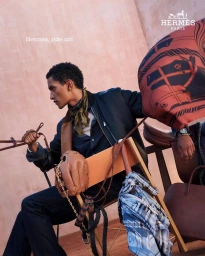 Man seated on a folding chair holding leather reins in an outdoor setting with Hermes branding.