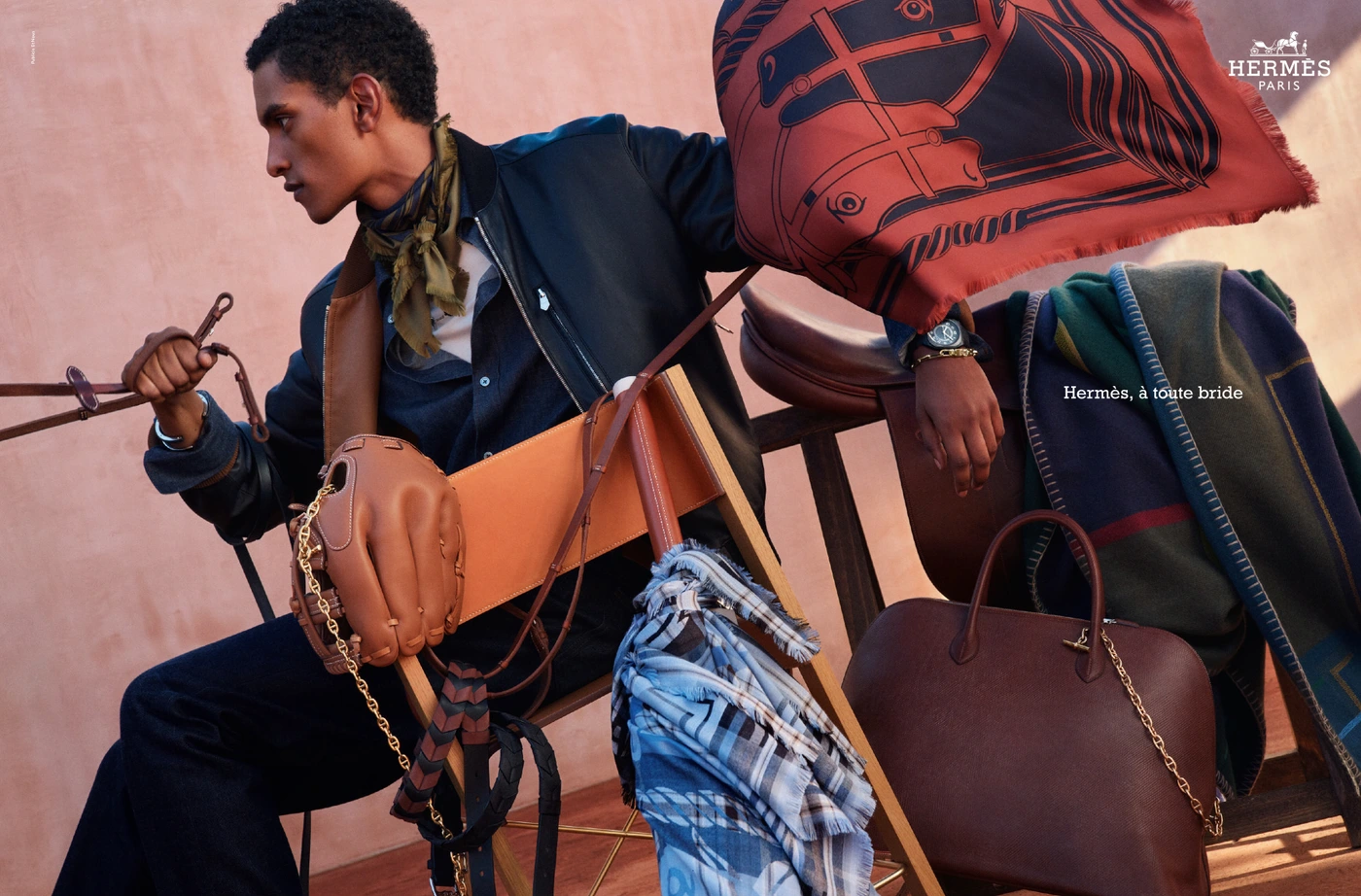 Man wearing dark jacket and silk scarf holding equestrian gear with Hermès leather bag and saddle in background