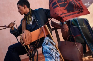 Man wearing dark jacket and silk scarf holding equestrian gear with Hermès leather bag and saddle in background