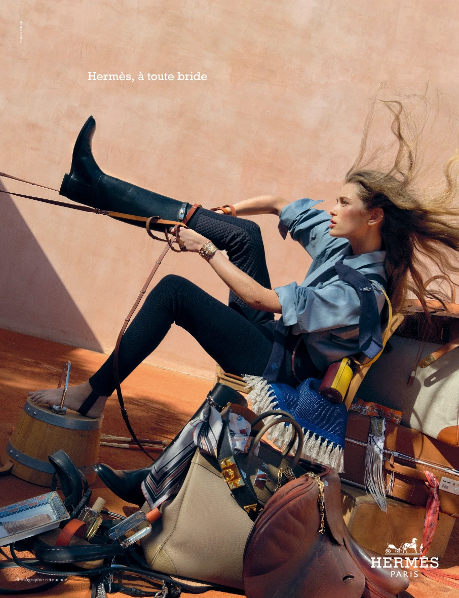 Model in blue shirt and black pants pulling on a tall riding boot, surrounded by Hermes luggage, boots, and leather saddles.