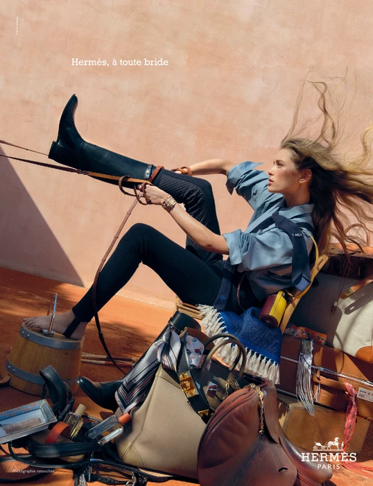 Model in blue shirt and black pants pulling on a tall riding boot, surrounded by Hermes luggage, boots, and leather saddles.