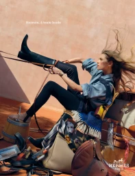 Model in blue shirt and black pants pulling on a tall riding boot, surrounded by Hermes luggage, boots, and leather saddles.
