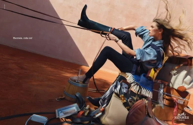 Model in blue shirt and tall black boots leaning back outdoors with wind in her hair amid leather bags and equestrian equipment