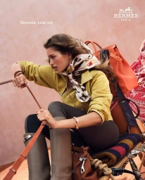 Woman in a yellow sweater and silk scarf holding reins against a textured terracotta wall with Hermes luggage