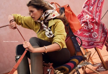 Model wearing yellow jacket, silk scarf, and tall boots holding leather reins against a pink wall with Hermes accessories.