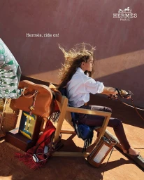 Model seated on a wooden chair holding leather reins against a warm textured pink wall for Hermes Ride On campaign