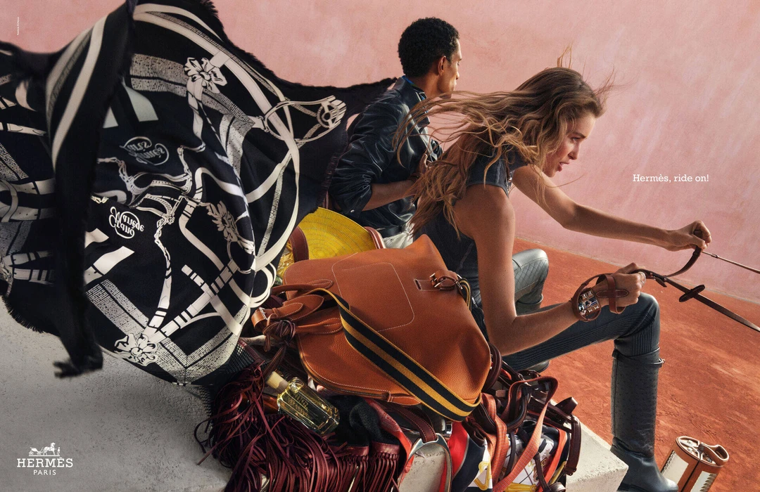 Models posing with Hermes leather bags and a large black and white patterned silk scarf outdoors against a pink wall