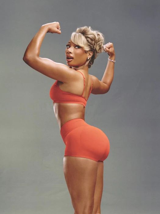 Megan Thee Stallion flexing biceps in an orange sports bra and matching shorts against a neutral grey studio background.