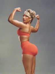 Megan Thee Stallion flexing biceps in an orange sports bra and matching shorts against a neutral grey studio background.