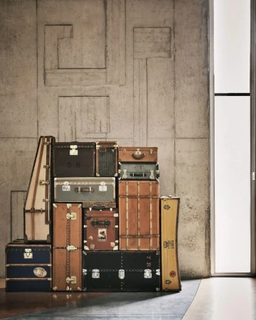 Stack of vintage Louis Vuitton trunks and suitcases against a grey concrete wall with a modern window, creating a travel-themed display.