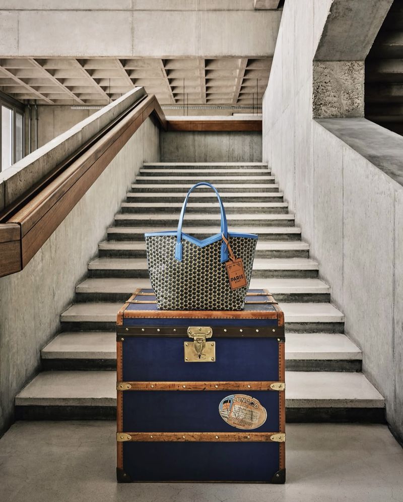 Tote bag and vintage trunk on staircase. Tan, black and blue tote bag is on top of a navy-blue wooden trunk with a staircase background.