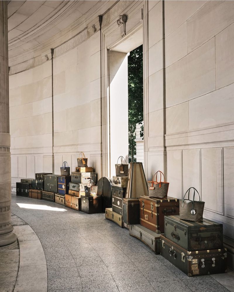 Stacked vintage luggage and tote bags create a display in a colonnade, casting shadows in the architectural space.