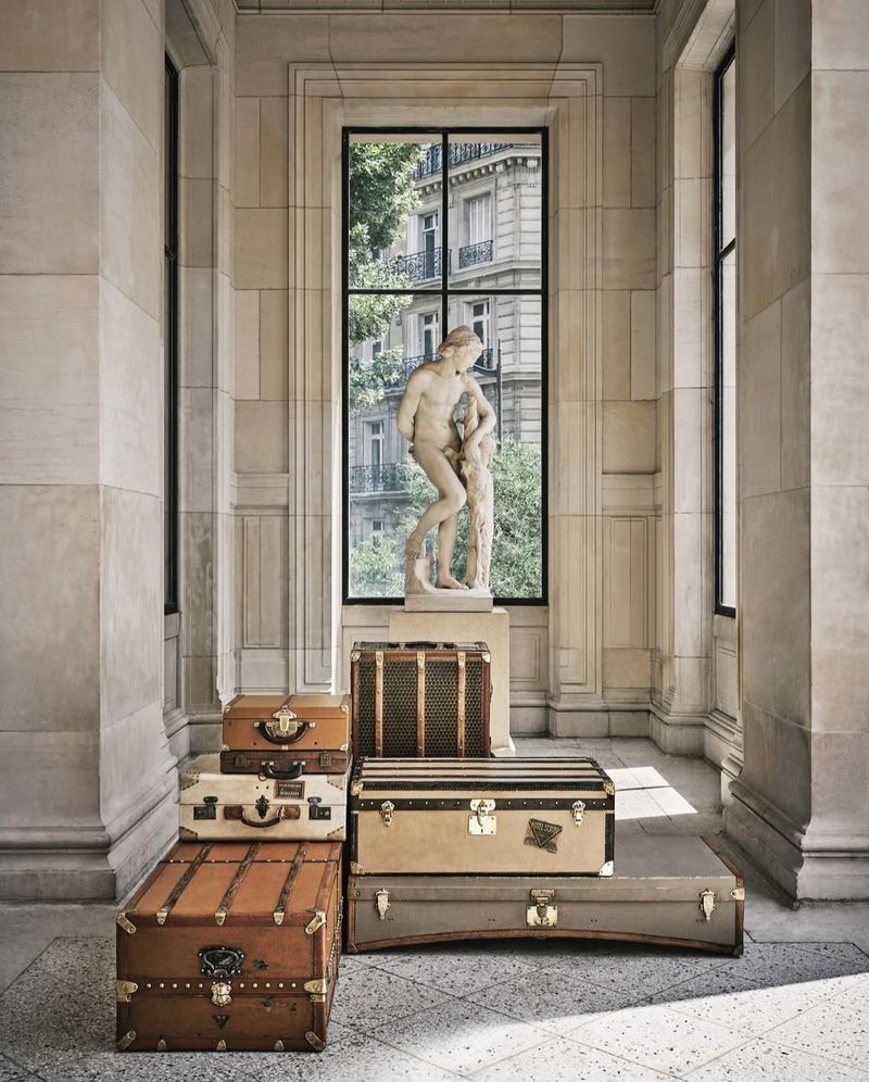 Stacked vintage luggage trunks in front of a window with a sculpture of a nude man in an indoor stone setting with natural light.