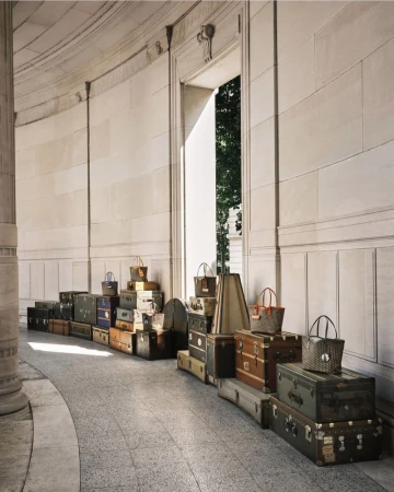 Vintage travel trunks and Moynat designer tote bags displayed along a curved stone architectural hallway with columns.