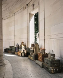 Vintage travel trunks and Moynat designer tote bags displayed along a curved stone architectural hallway with columns.