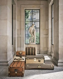 Vintage Moynat luxury luggage trunks stacked in a neoclassical stone hall with a classical statue and tall window.