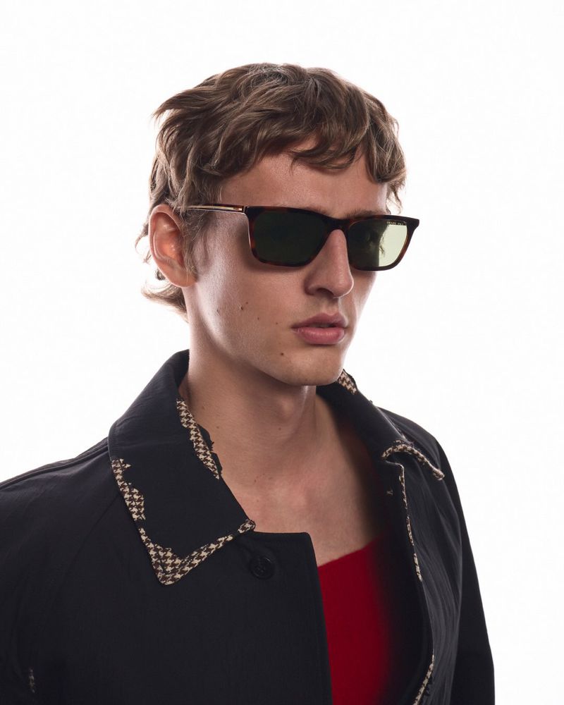 Close-up portrait of a male model wearing tortoiseshell sunglasses and a dark jacket with patterned collar against a plain white background.